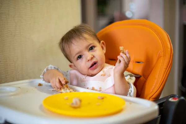 Lindo bebé en un asiento de niño naranja inclinó su cabeza y mira a la cámara, sosteniendo un pastel en sus manos. migas y un plato naranja sobre la mesa. primer plano, vista frontal, enfoque suave, fondo borroso — Foto de Stock
