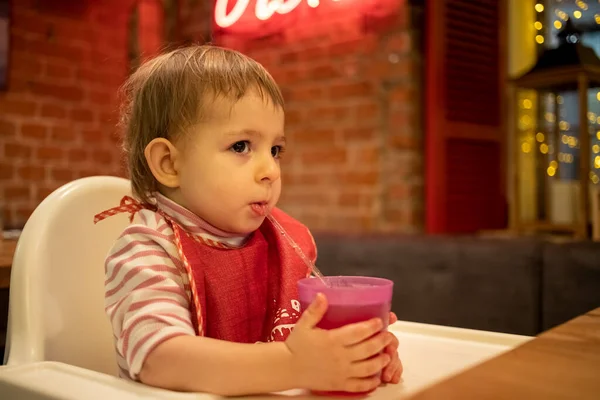 Un niño lindo se sienta en una silla alta, sostiene un vaso de jugo con las manos y bebe de una pajita. en el fondo un acogedor interior de restaurante con paredes de ladrillo — Foto de Stock