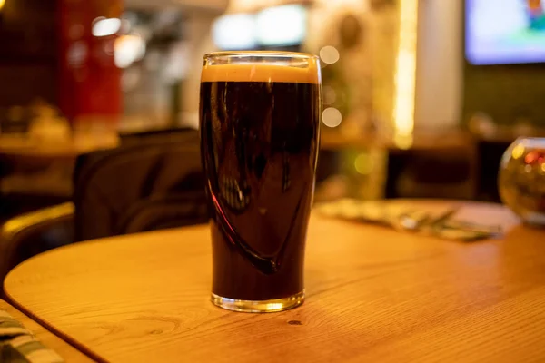 pint glass of dark stout beer with foam stands on a wooden table in a bar