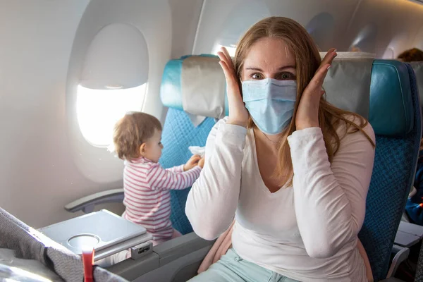 Retrato de una madre joven y asustada en una silla de avión con una máscara respiratoria médica que mira a la cámara horrorizada por el coronavirus Covid-19. un lindo bebé está sentado a su lado — Foto de Stock