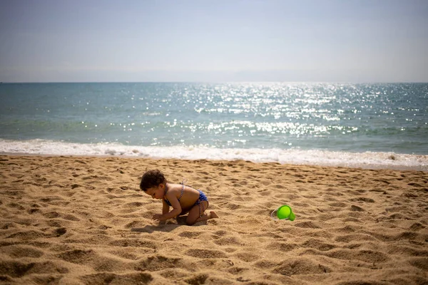 Mała słodka dziewczynka maluch w niebieskim stroju kąpielowym na piaszczystej plaży bawi się w piasek na tle fal morskich w ciepły słoneczny dzień — Zdjęcie stockowe