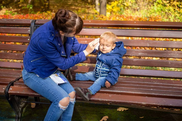 young beautiful mom in blue clothes on a bench in the park wipes the babys dirty face with a napkin. baby is in blue clothes. close up, soft focus, on the background of trees in blur