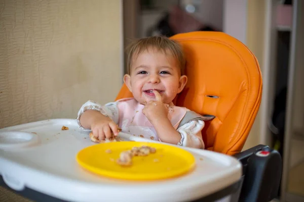 Lindo bebé en un asiento de bebé naranja mira la cámara riendo mostrando la mano en los dientes.Los dientes han crecido. migas y un plato naranja con un pastel en la mesa . — Foto de Stock