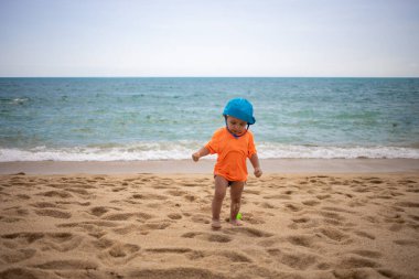 Mavi şapkalı, turuncu tişörtlü, kumsalda yürüyen bebek. Ön Manzara. Karantina, seyahat özgürlüğü, açık sınırlar