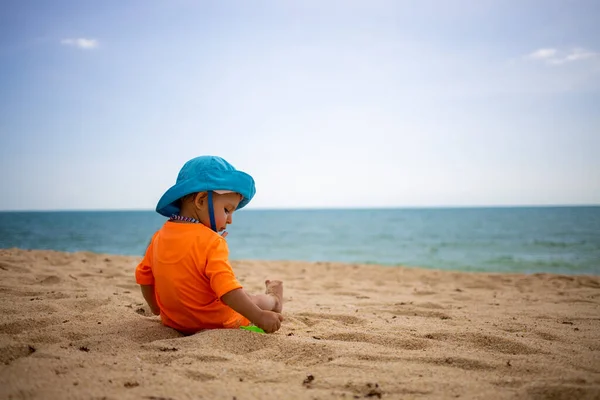 Szczęśliwy maluch w niebieskim kapeluszu i pomarańczowym t-sircie bawi się piaskiem na tropikalnej plaży i czepia się piasku. Morze jest na bachgound — Zdjęcie stockowe