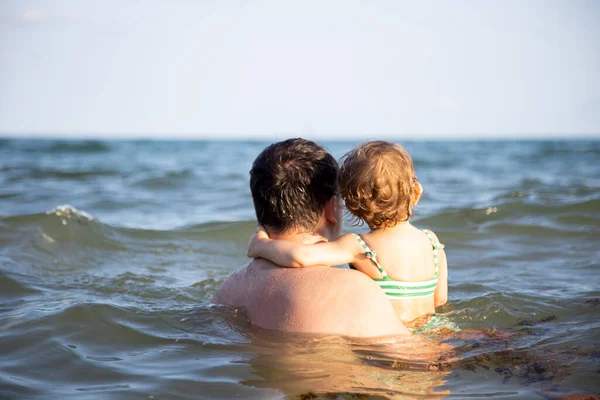 お父さんは海の水の中にかわいい幼児の娘を持っていて地平線の後ろの景色を見て — ストック写真