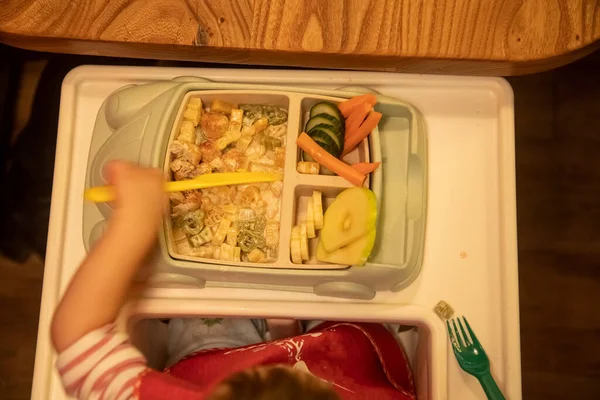 Vista superior, el niño se sienta frente a un plato para niños lleno de deliciosa comida deliciosa y corta la comida con un cuchillo de plástico para niños. enfoque suave — Foto de Stock