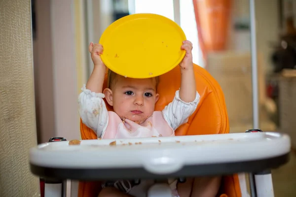 Un lindo bebé caucásico se sienta en una silla de bebé naranja con una mesa después de una comida y juega con un plato de plástico naranja. primer plano, vista frontal, enfoque suave — Foto de Stock