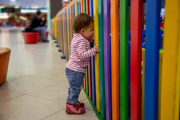 Un niño pequeño y lindo está de pie en la valla de colores de una sala de juegos para niños se ve a través de ella triste y quiere jugar dentro. enfoque suave. primer plano, fondo borroso — Foto de Stock