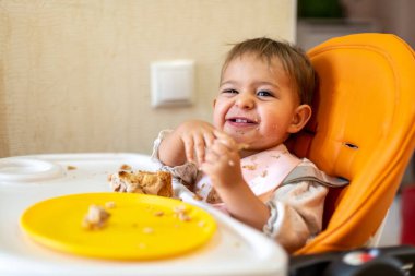 Şirin bebek, kirli elleri ve yüzü olan bir masası olan turuncu bir bebek sandalyesinde oturur, kameraya bakar ve güler. Masada kırıntılar ve turuncu bir tabak var. yakın plan, kenar görünüm, yumuşak odak, bulanık arkaplan