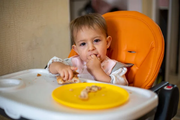 Lindo bebé en una silla de bebé naranja mira a la cámara y disfruta comiendo el pastel con las manos. migas y un plato naranja sobre la mesa. primer plano, vista frontal, enfoque suave, fondo borroso . — Foto de Stock