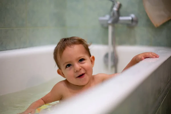 Крихітне кавказьке дитя приймає ванну, грайливо посміхається і дивиться вгору, відпочиваючи на стороні ванни. Вода бризкає, на задньому плані зелена ванна розмивається. М'який фокус. — стокове фото