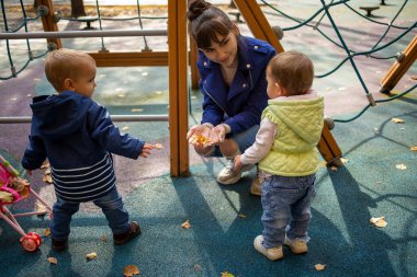 Mavi ceketli güzel bir anne, bir sonbahar parkında, mavi elbiseli ve gülümseyen 1 yaşındaki küçük tatlı oğlunun ve kızının önünde çömelmiş, ona sarı yaprakları gösteriyor.