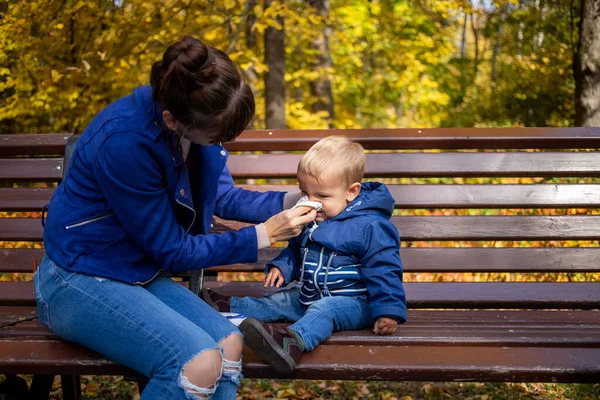 Autumn cold in children - concept. a young mother in blue clothes on a park bench wipes the nose of little baby who has a runny nose and blows nose. close-up, soft focus, on background trees in blur. — Stock Photo, Image