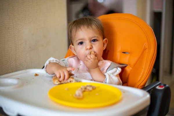 Lindo bebé en una silla de bebé naranja mira a la cámara y disfruta comiendo el pastel con las manos. migas y un plato naranja sobre la mesa. primer plano, vista frontal, enfoque suave, fondo borroso — Foto de Stock