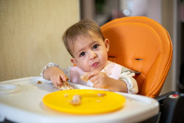Lindo bebé en un asiento de niño naranja inclinó su cabeza y mira a la cámara, sosteniendo un pastel en sus manos. migas y un plato naranja sobre la mesa. primer plano, vista frontal, enfoque suave, fondo borroso — Foto de Stock