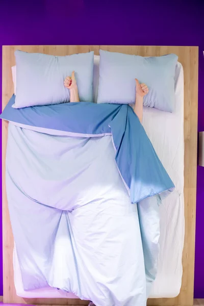 Sign of like thumbs up show two female hands lying on a pillow from under the blue covers in bed, top view, wooden bed