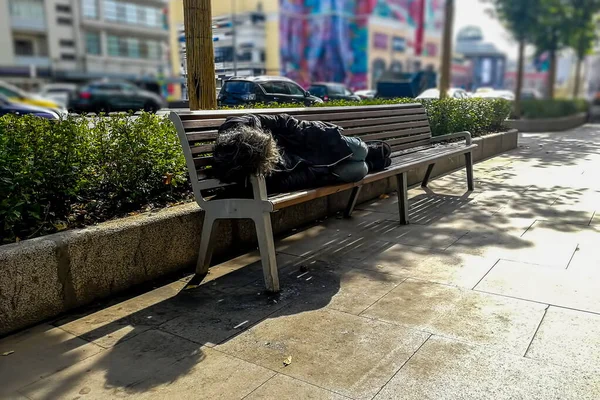 Homeless beggar dirty man sleeping on a bench under a tree on the street, in the background the city, traffic, shopping center and colorful houses in blur. soft focus, close up. — Stock Photo, Image