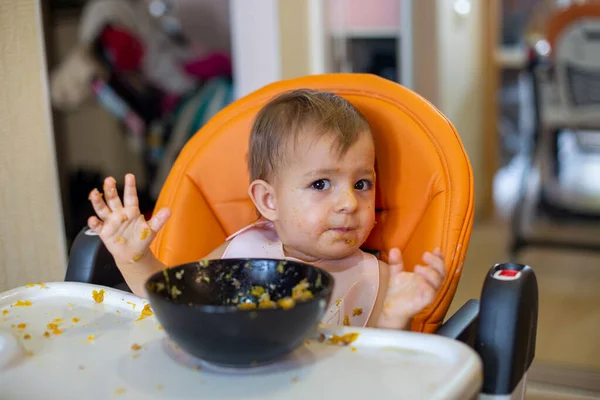 Linda niña en un asiento de niño naranja delante de un plato de comida arroja sus manos y mira a la cámara. por todas partes en migas de comida. primer plano, vista frontal, enfoque suave, fondo borroso — Foto de Stock