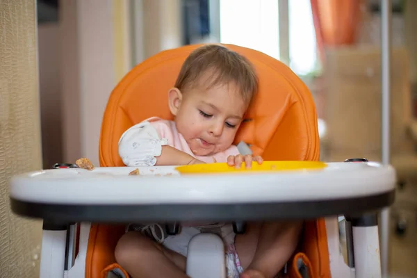 Lindo bebé caucásico se sienta en una silla de bebé naranja después de comer cava en el delantal, en las migajas de mesa y un plato de plástico naranja. primer plano, vista frontal, enfoque suave, fondo borroso — Foto de Stock