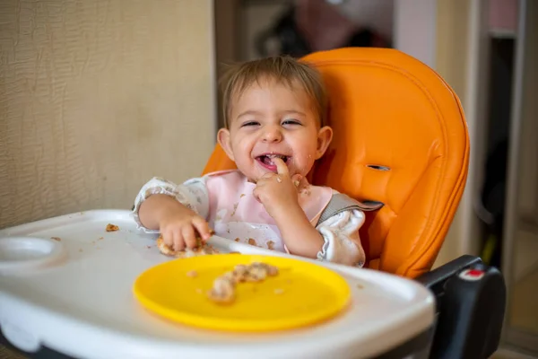 Lindo bebé en un asiento de niño naranja mira a la cámara riendo mostrando la mano en los dientes. Los dientes han crecido. migas y un plato naranja con un pastel en la mesa. primer plano, vista frontal, enfoque suave — Foto de Stock