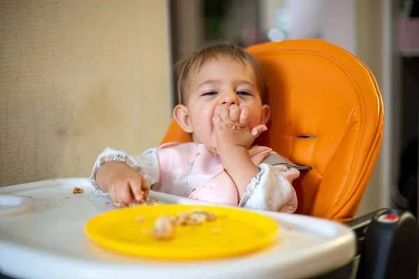 Lindo bebé en una silla de bebé naranja mira a la cámara y disfruta comiendo el pastel con las manos. migas y un plato naranja sobre la mesa. primer plano, vista frontal, enfoque suave, fondo borroso — Foto de Stock