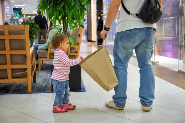 Venerdì nero concetto, shopping con i bambini. piccola ragazza carina con papà tiene un sacchetto di carta artigianale per lo shopping e guarda il padre. primo piano, soft focus, sullo sfondo un centro commerciale in sfocatura — Foto Stock