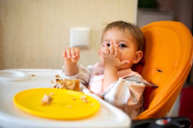 Şirin bebek, kirli elleri ve yüzü olan bir masası olan turuncu bebek koltuğunda oturur kameraya bakar ve hava öpücüğü gönderir. Masada kırıntılar ve turuncu bir tabak var. Yakın çekim, yan görünüm, yumuşak odaklanma, 