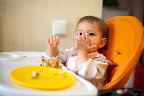 Lindo bebé se sienta en una silla de bebé naranja con una mesa con las manos y la cara sucias, mira en la cámara y enviar un beso de aire. sobre la mesa hay migajas y un plato de color naranja. primer plano, vista lateral, enfoque suave , — Foto de Stock