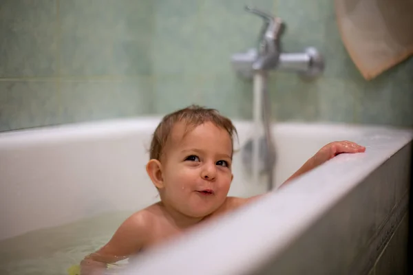 Крихітне кавказьке дитя приймає ванну, усміхається несміливо і дивиться вгору, відпочиваючи на стороні ванни. Вода бризкає, на задньому плані зелена ванна розмивається. М'який фокус. — стокове фото