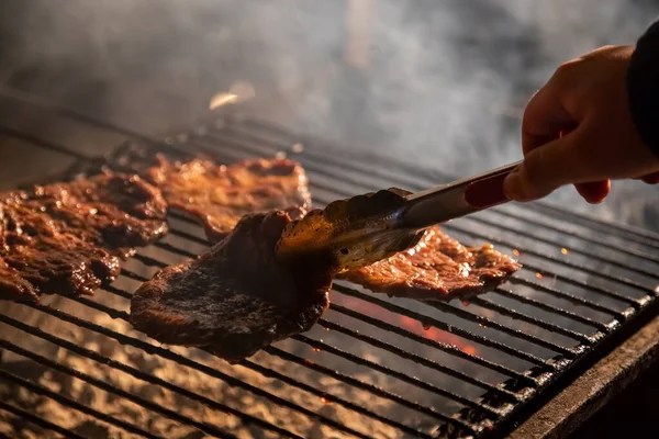 Abends gibt es einen Grill, auf dem leckere saftige Steaks über offenem Feuer gegrillt werden, eine Hand hält eine Zange und dreht das Fleisch um. Nahaufnahme, weicher Fokus. Rauch wird hervorgehoben. — Stockfoto