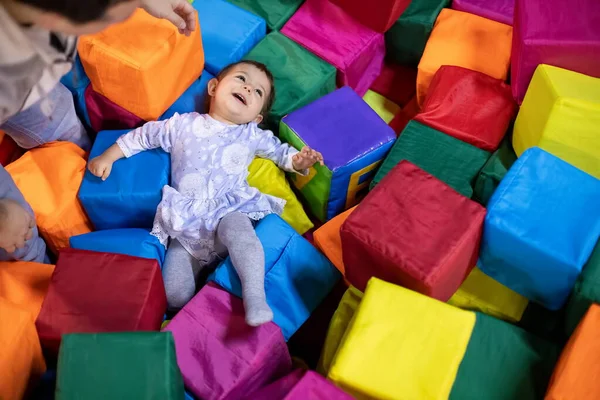 Little cute toddler girl in a dress lies with smile and happy laugh in the pool with cubes her fater stays near.. playground in the childrens play center. top view Stock Picture