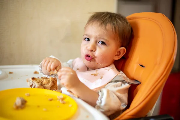 Un bebé lindo está sentado en un asiento de niño naranja con una mesa con las manos y la cara sucias, mirando a la cámara y haciendo muecas. hay migajas y un plato naranja en la mesa . — Foto de Stock