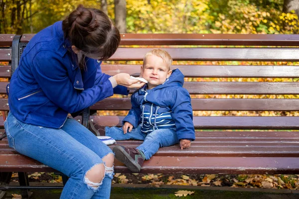 Pequeno bebê bonito em roupas azuis senta-se em um banco no parque, olha para a câmera seriamente, enquanto a mãe limpa seu rosto sujo com um guardanapo. close-up, no fundo são árvores de outono em borrão . — Fotografia de Stock