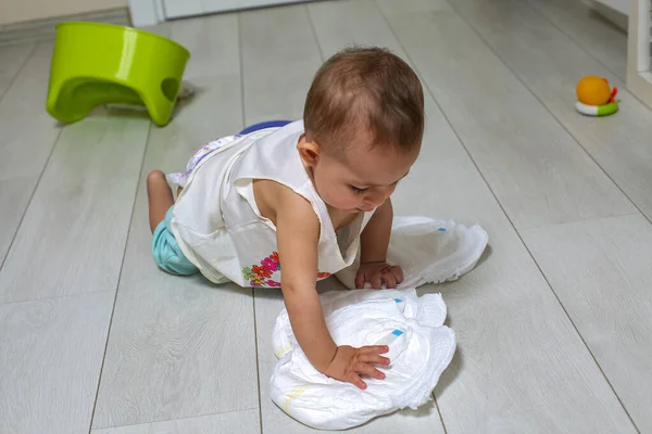 Un pequeño bebé lindo juega con pañales nuevos y limpios en el suelo. en el fondo una olla de cámara y juguetes en borroso. El concepto de aprender a usar el pote de la cámara . — Foto de Stock