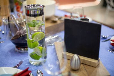 Mavi masa örtüsü ile servis edilen bir masa, çelik aletler ve tuzluk, kırmızı peçeteli beyaz ve çıplak bir tabak, limon suyu dolu bir sürahi ve siyah bir plaka var..