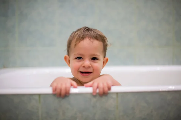 Миле кавказьке немовля визирає з ванни, кладе руки на край ванни і дивиться на камеру з посмішкою і сміхом. На задньому плані - зелена ванна в тумані. М'який фокус.. — стокове фото
