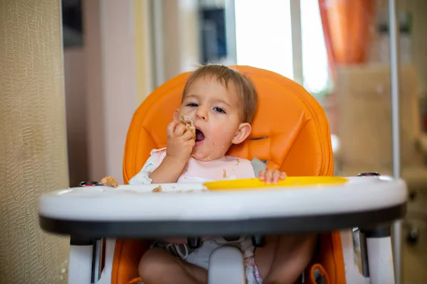 Lindo bebé caucásico sentado en una silla de bebé naranja comiendo un pastel con las manos de un plato de plástico naranja y mirando a la cámara. primer plano, vista frontal, enfoque suave, fondo borroso — Foto de Stock