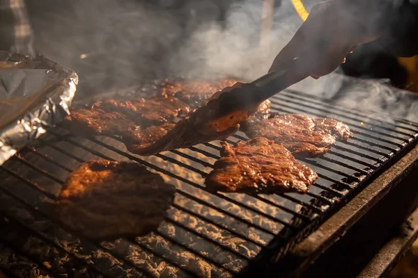 Večer gril na grilu, na kterém se grilují chutné šťavnaté steaky nad otevřeným ohněm, ruka drží kleště a převrací maso. zblízka, měkké soustředění. kouř je zvýrazněn — Stock fotografie