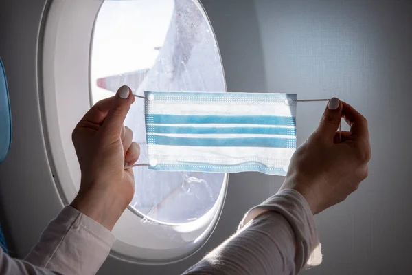 Concept flights to asia coronavirus epidemic. close-up female hands holding a medical mask against the background of an airplane porthole — Stock Photo, Image