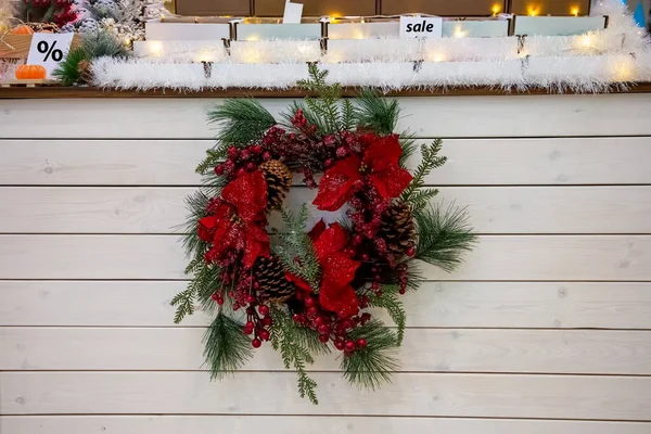 A beautiful Christmas wreath of coniferous fir branches, red berries and flowers in hoarfrost hanging on background of light wooden counter. upstairs decorated display case with inscription sale and — Stock Photo, Image