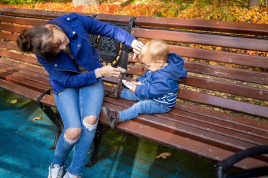 Anne parkta bir bankta bebek yüzünü peçeteyle siliyor. Bebek yürürken kirlenmiş. Yakın plan, yumuşak odak, bulanık ağaçların arka plan karşı