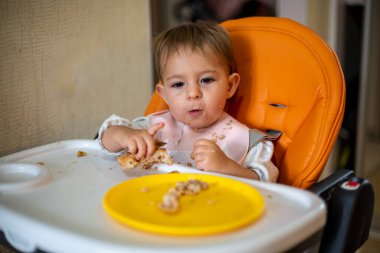 Turuncu bebek koltuğundaki şirin bebek bir dilim pasta seçiyor. Kırıntılar ve masada turta olan turuncu bir tabak. yakın plan, ön görüş, yumuşak odaklanma, bulanık arkaplan