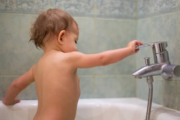 Küçük tatlı bir çocuk, banyoda banyo yaparken muslukla oynar, musluğu açar ve kapatır. Yeşil bir banyonun arka planına yakın plan, yumuşak odak