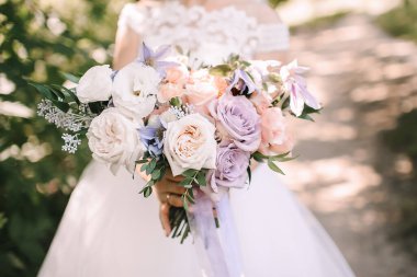 Gelin buketine yakın plan güller, karanfiller beyaz, pembe, menekşe, lavanta, şeftali rengi. Dantelli beyaz elbiseli gelin elinde bir buket tutuyor.