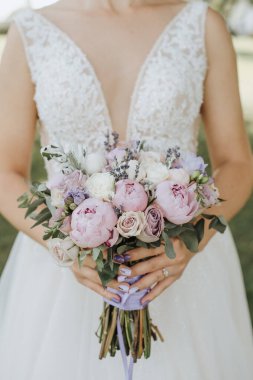 Bir buket şakayık, gül, karanfil, beyaz, pembe, menekşe, lavanta rengi. Dantelli beyaz elbiseli gelin elinde bir buket tutuyor.