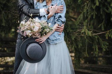 Kot ceketli gelin elinde beyaz ve mor gözlüklü bir şapka ve bir buket tutuyor. Damat ona arkadan sarılıyor.