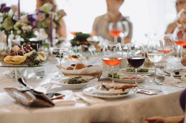 Ziyafet. Masayı beyaz bir masa örtüsüyle örtün. Masanın üzerinde şarap bardakları ve yemekli tabaklar var. Bir sürü insanın arasında.