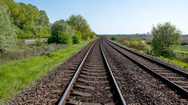 16.9 ekin paralel tren yolu uzaklığa doğru ilerliyor. Etrafı yemyeşil ağaçlarla çevrili. Mavi gökyüzünün altında çalılıklar var.