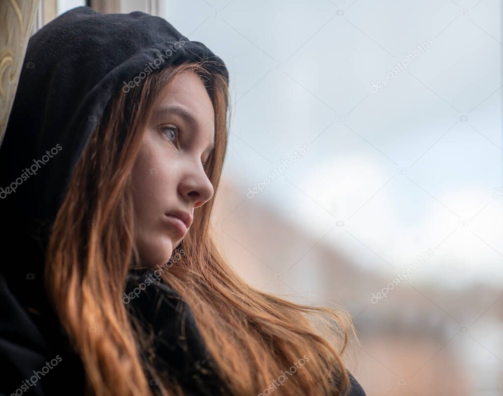 Low angle sad teen girl in black hoodie resting near window and looking depressed isolated teenager  maybe suffering from child abuse 